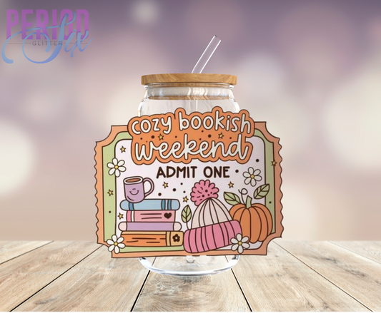 A clear glass jar with a label that reads 'Cozy Bookish Weekend ADMIT ONE' surrounded by decorative elements like books, a mug, and a pumpkin on a wooden surface.